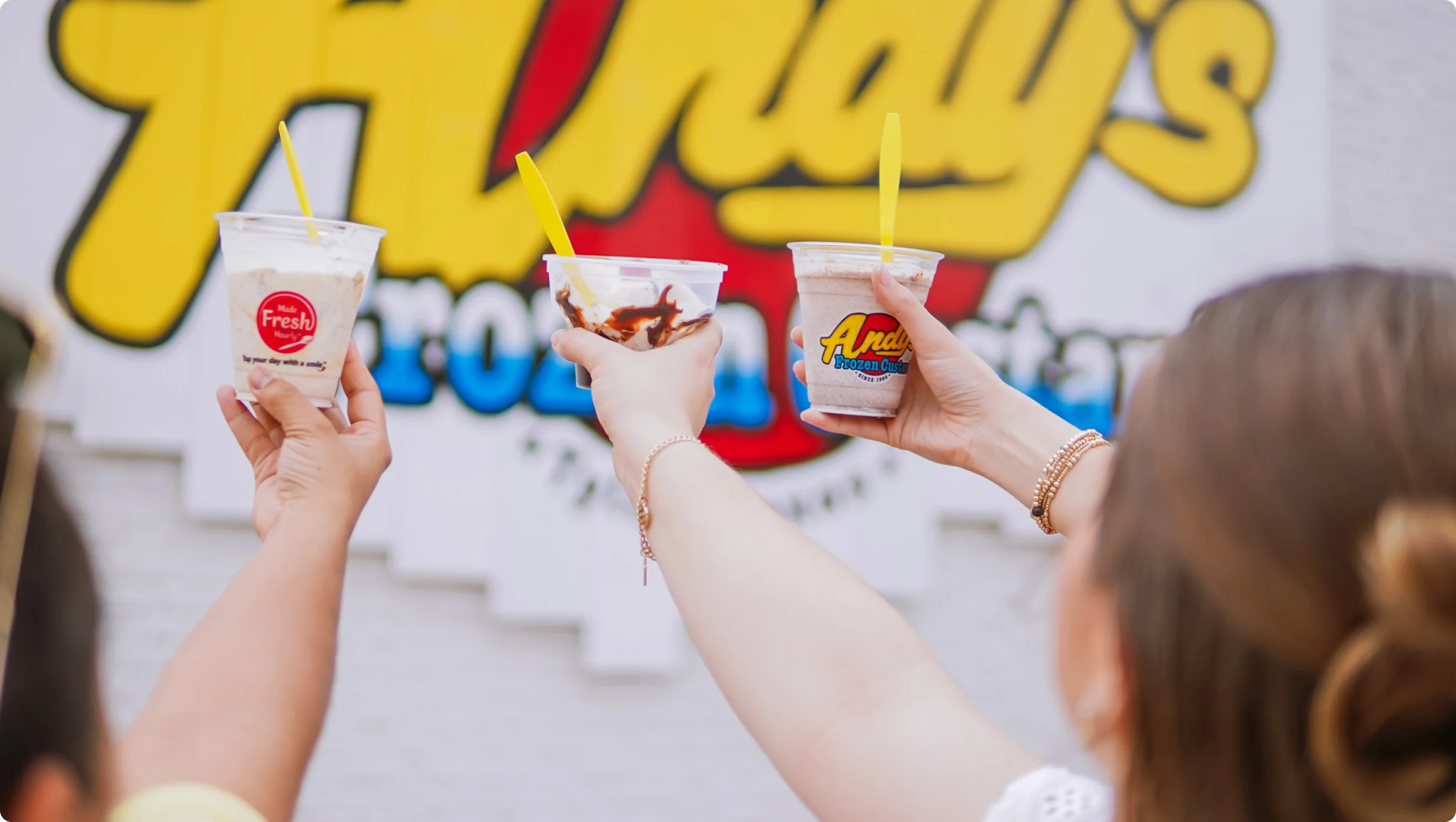 Friends raising Andy’s Frozen Custard concretes in front of the Andy’s sign